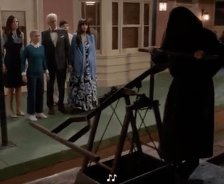 a still image from the TV show The Good Place in which a hooded ominous figure is arriving via a train-track handcart, to an audience of shocked and concerned characters